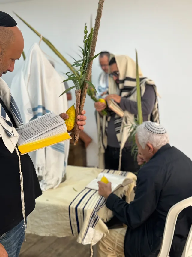 מתפלל מבוגר יושב עם אתרוג, ולידו מתפללים עם לולב