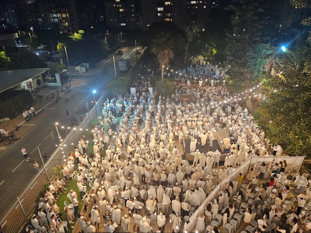 צילום אוויר לילי של מאות מתפללים עטופים בטליתות לבנות, מתפללים בשטח פתוח בין בנייני מגורים, מוארים בשרשרות נורות.