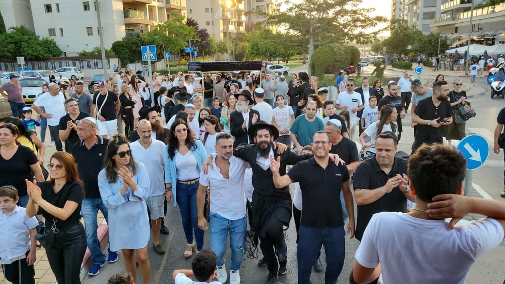תהלוכת הכנסת ספר תורה ברחוב בשכונה, עשרות תושבים מכל הסוגים צועדים ומוחאים כפיים יחד לאור שקיעה.